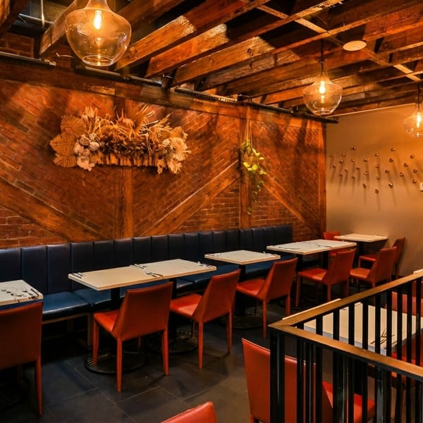 Cozy restaurant interior with exposed brick, wooden beams, blue banquettes, red chairs, and hanging lights.