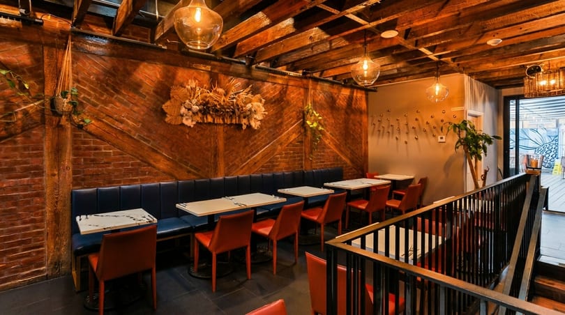 Cozy restaurant interior with exposed brick, wooden beams, blue banquettes, red chairs, and hanging lights.