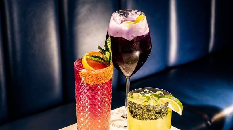 Three colorful cocktails with garnishes, including citrus and mint, against a dark blue background.