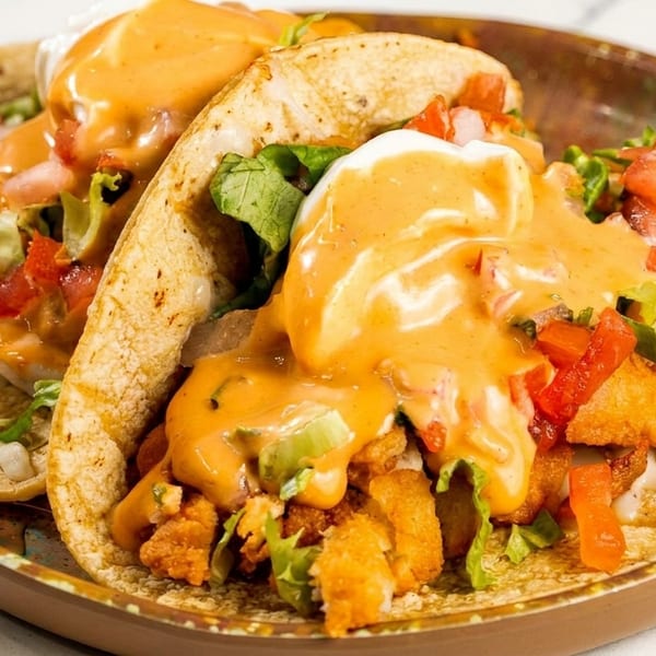 Two crispy chicken tacos with lettuce, tomatoes, and creamy orange sauce on a brown plate.