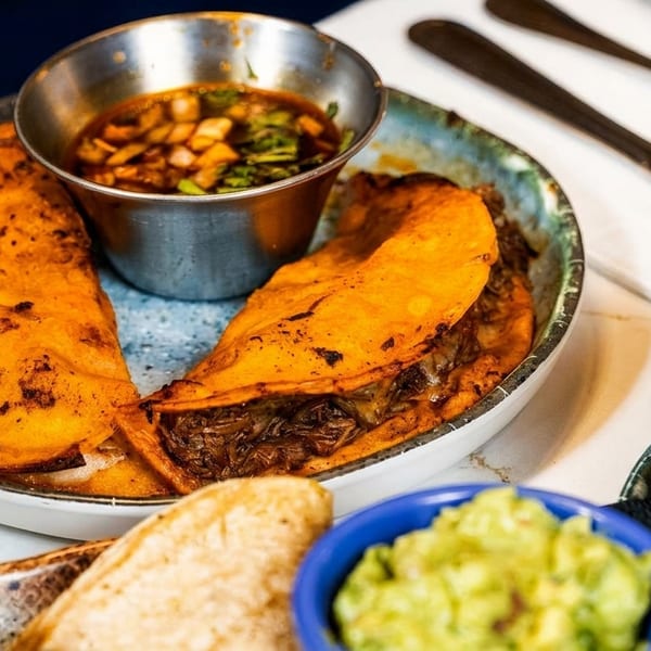 Two birria tacos with consommé, guacamole, and another dip on a marble table.