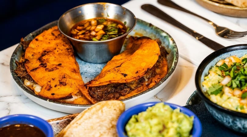 Two birria tacos with consommé, guacamole, and another dip on a marble table.