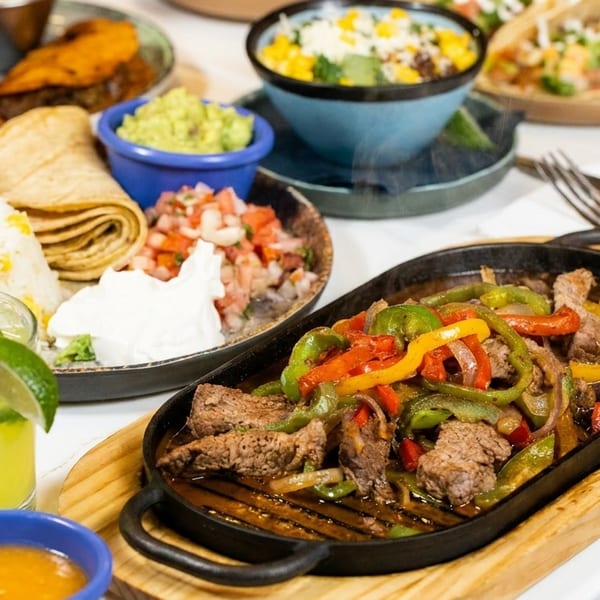 Sizzling beef fajitas with colorful peppers, rice, tortillas, guacamole, and a margarita on a table.