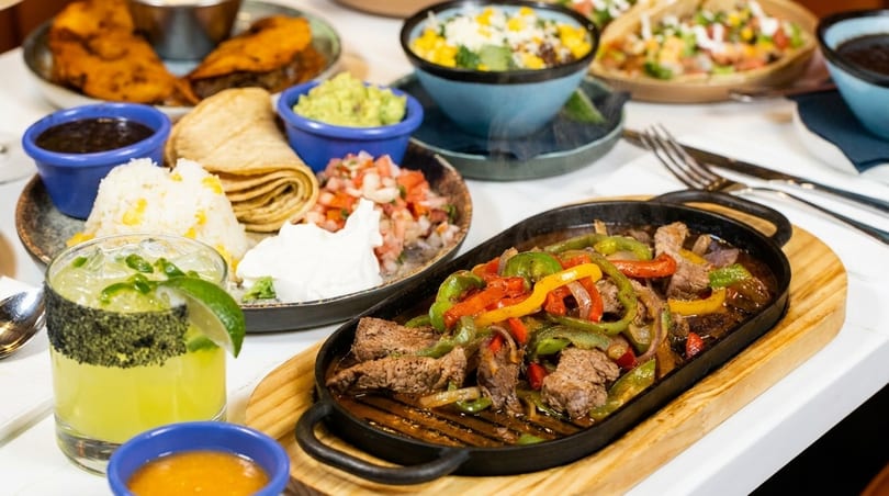 Sizzling beef fajitas with colorful peppers, rice, tortillas, guacamole, and a margarita on a table.