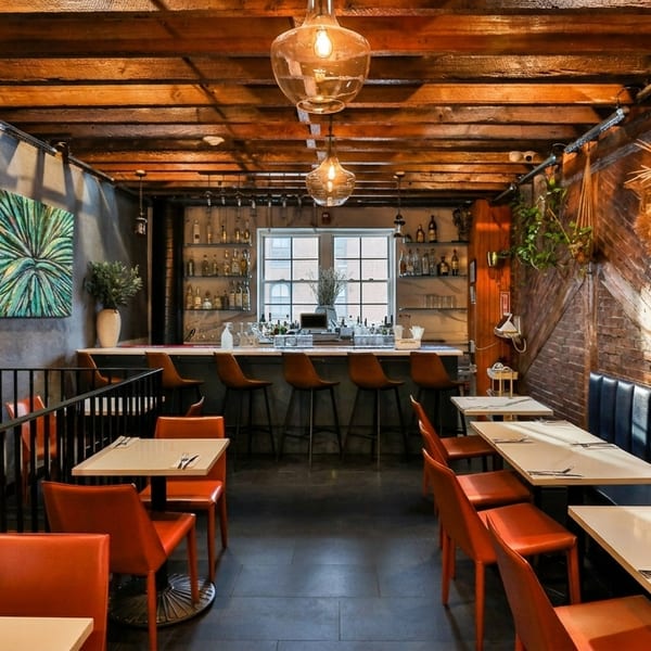 A restaurant interior with a bar, orange chairs, blue banquette seating, and exposed brick and wood.