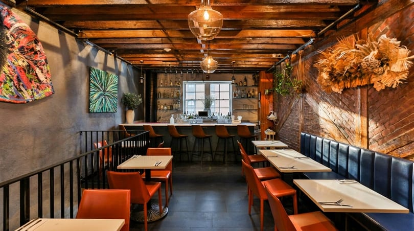 A restaurant interior with a bar, orange chairs, blue banquette seating, and exposed brick and wood.