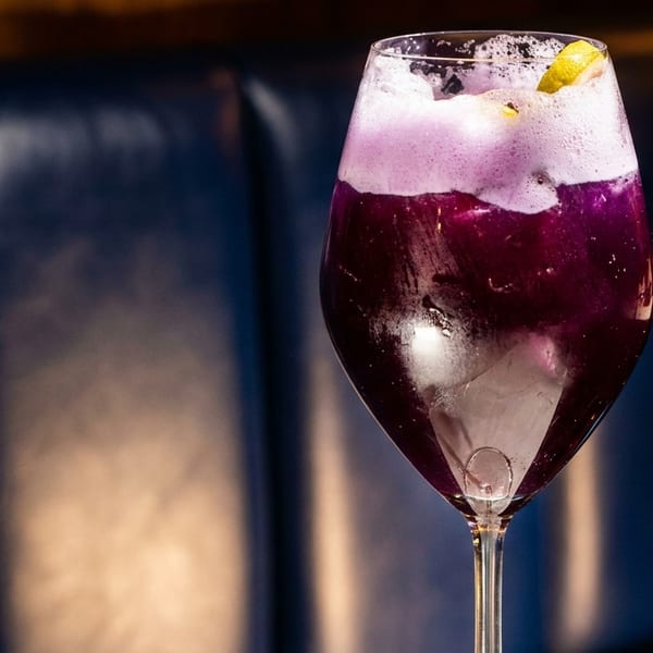 Purple cocktail with white foam and a lemon slice in a wine glass against a dark background.