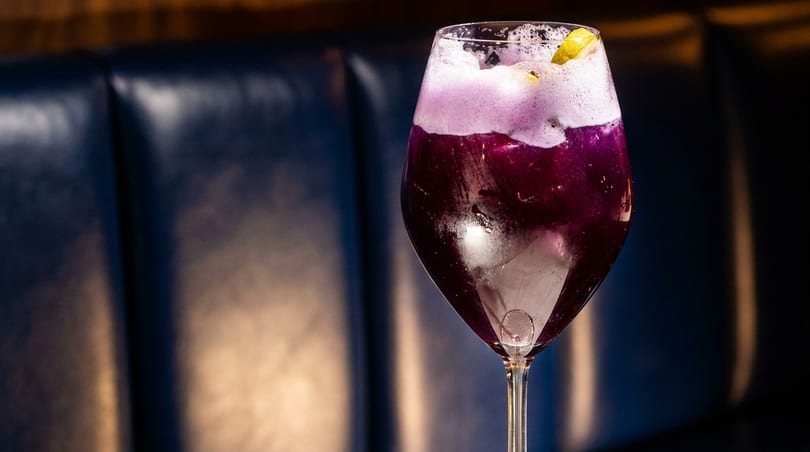 Purple cocktail with white foam and a lemon slice in a wine glass against a dark background.