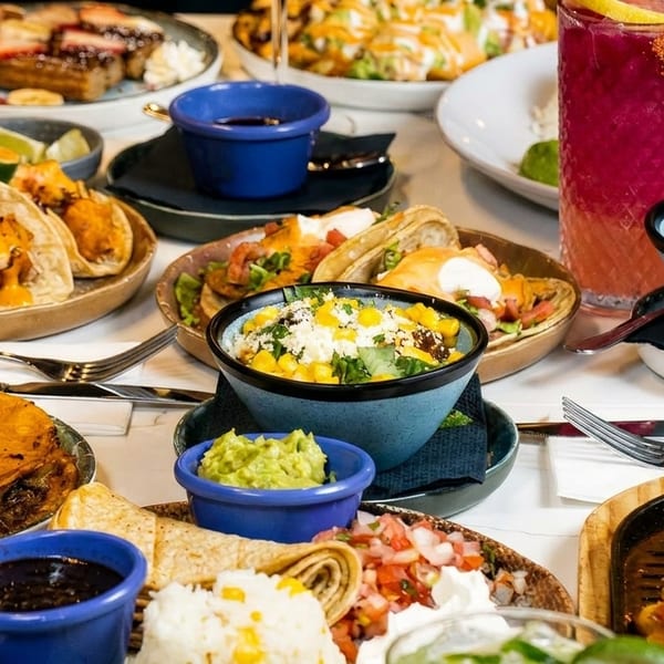 A table laden with various Mexican dishes, including tacos, fajitas, elote, and drinks.