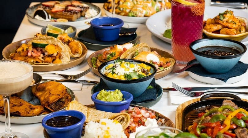 A table laden with various Mexican dishes, including tacos, fajitas, elote, and drinks.